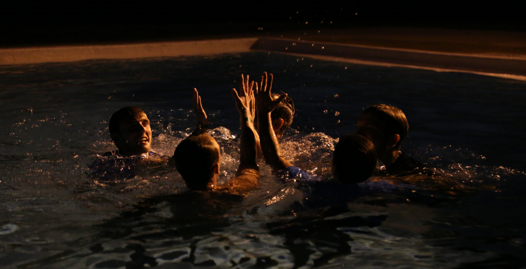 Group of people in a dark pool, hands raised up as water splashes around them in a nighttime celebration.
