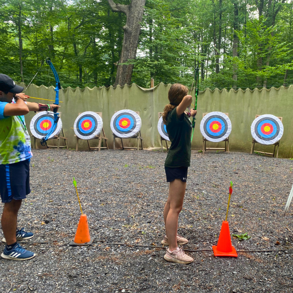 Person drawing a green bow at an outdoor archery range with targets set against a wooden fence and trees behind.