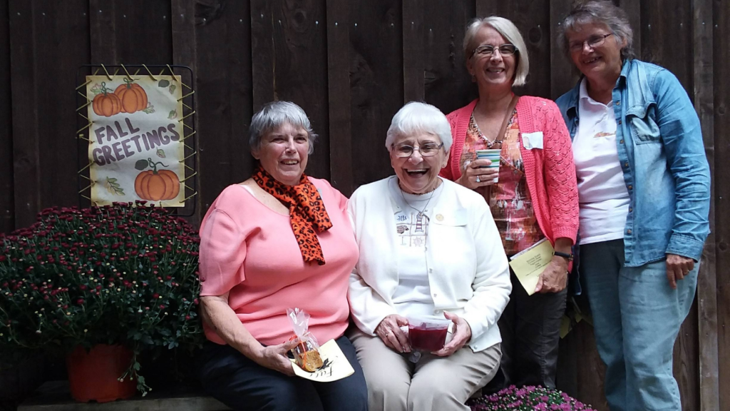 Four smiling women posing outdoors by a wooden fence, fall sign and potted mums on the left side.