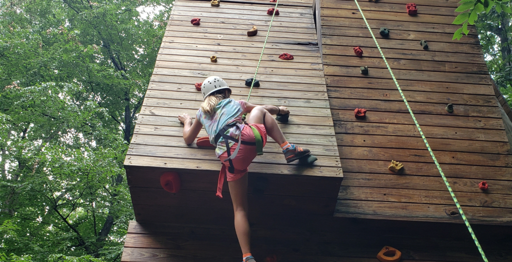 Child wearing a helmet climbs a tall wooden outdoor climbing wall with colorful holds and a safety rope.