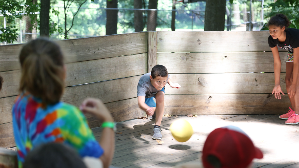 Children playing dodgeball inside a wooden outdoor arena; a boy in a gray shirt throws a yellow ball toward teammates.