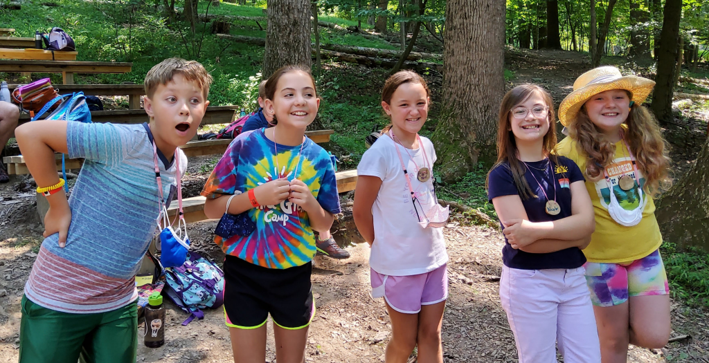 Group of five children at a campsite posing and smiling, wearing colorful clothing and lanyards in a forested area with picnic benches nearby.