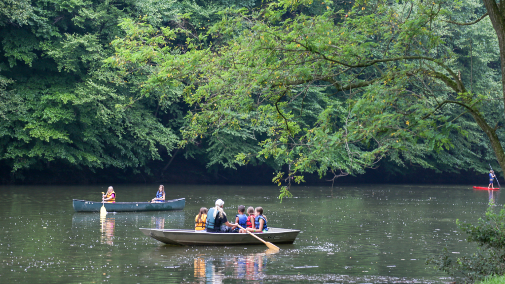 Children in life jackets paddling a canoe on a calm lake, with dense green trees along the shore.