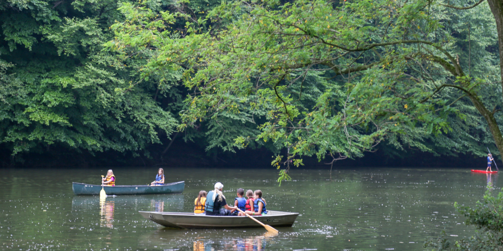 Children in life jackets paddling a canoe on a calm lake, with dense green trees along the shore.